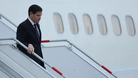 Getty Images Marco Rubio walks off a plane holding a rail wearing a suit